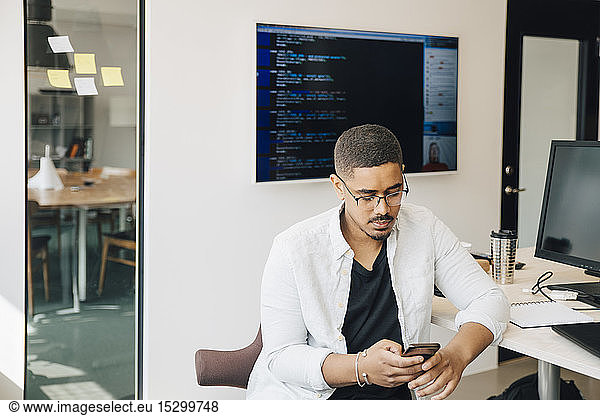 Male computer hacker using smart phone while sitting at desk in creative office