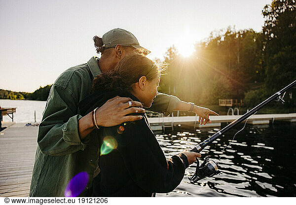 Male counselor gesturing by girl fishing on jetty near lake at summer camp