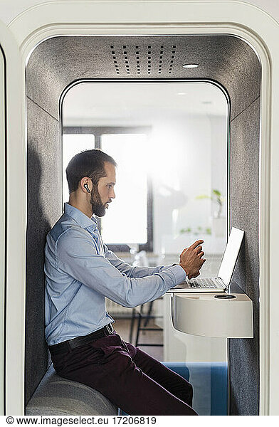 Male entrepreneur attending meeting on laptop in telephone booth at office