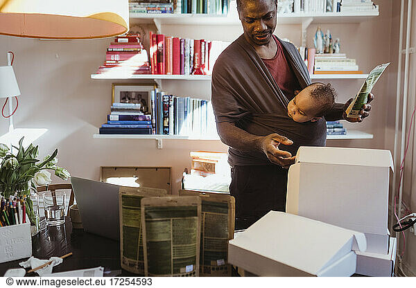 Male entrepreneur examining boxes while carrying baby boy in carrier at home office