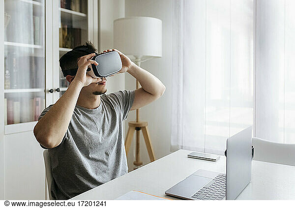 Male entrepreneur holding Virtual reality headset while sitting with laptop on desk at home office