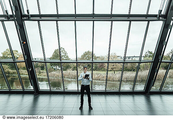Male entrepreneur using digital tablet while using digital tablet against glass in office corridor