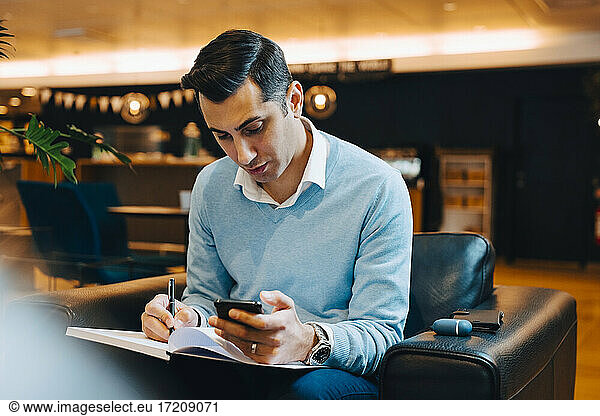 Male entrepreneur with smart phone writing in book at office