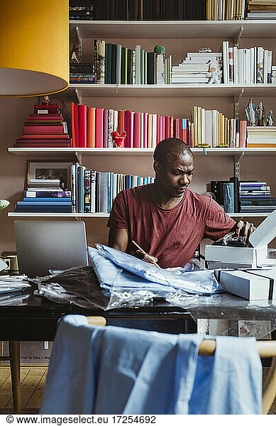 Male entrepreneur writing while examining shirt at home office