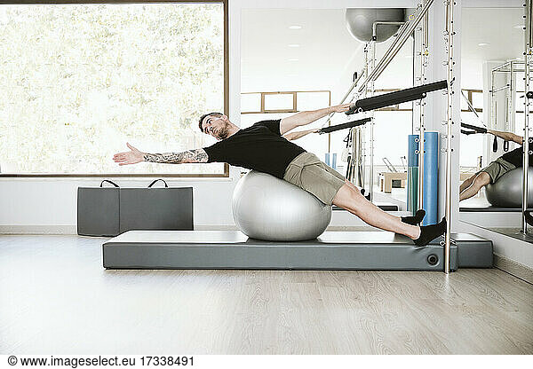 Male fitness instructor stretching arms on fitness ball in pilates studio