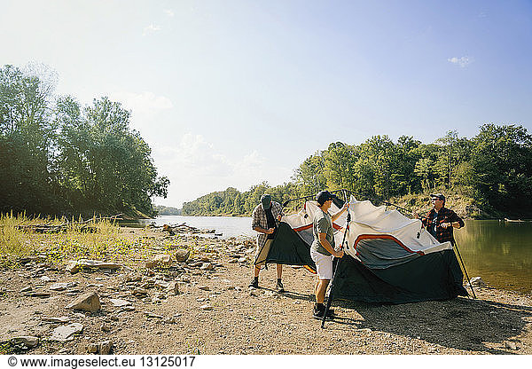 Male friends assembling tent at campsite by lake against sky