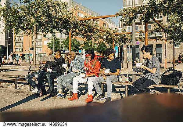 Male friends eating take out food while sitting side by side on bench in city