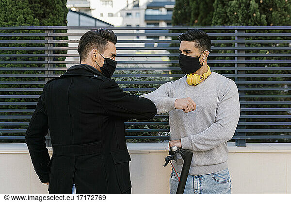 Male friends greeting each other by giving elbow bump wearing protective face mask against metal fence during COVID-19