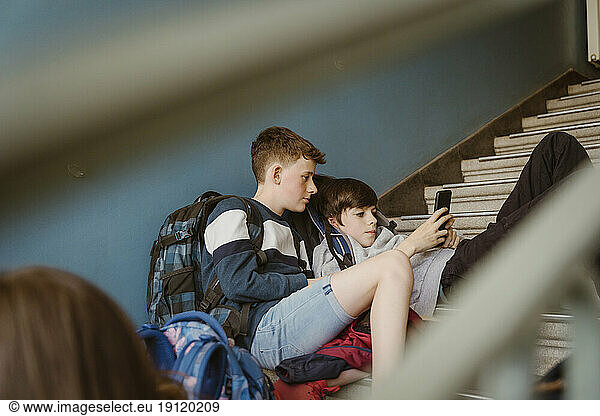 Male friends sharing smart phone while sitting on staircase of school building