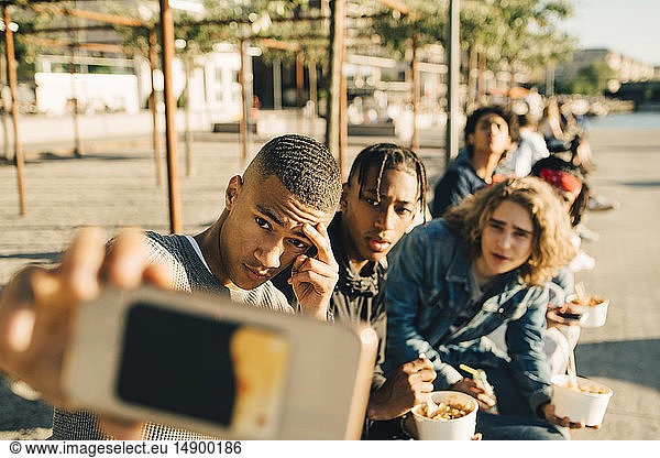 Male friends taking selfie while eating take out food on street in city
