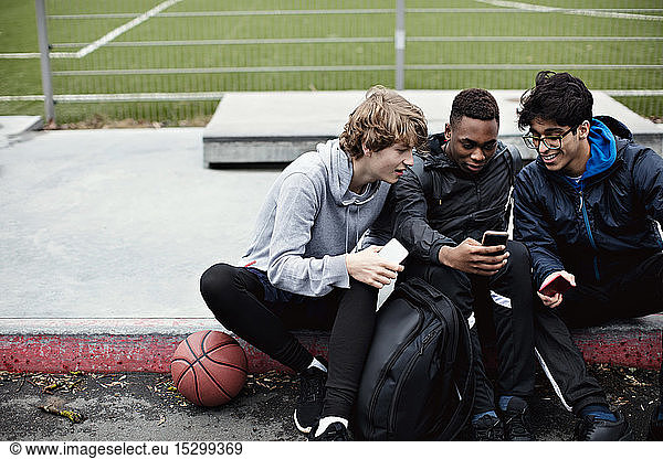 Male friends using mobile phones while sitting on sidewalk after basketball practice during winter