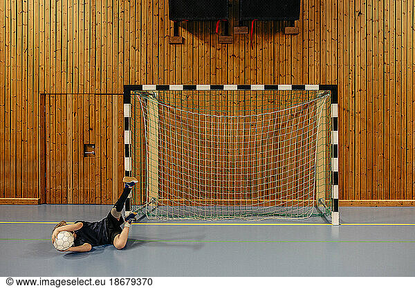Male goalie with disabled leg lying on floor holding football while playing at sports court