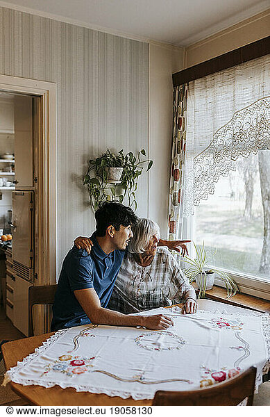 Male healthcare worker gesturing while sitting with senior woman near window at home