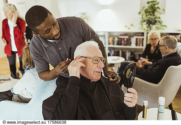 Male healthcare worker talking with senior man while cutting hair in retirement home