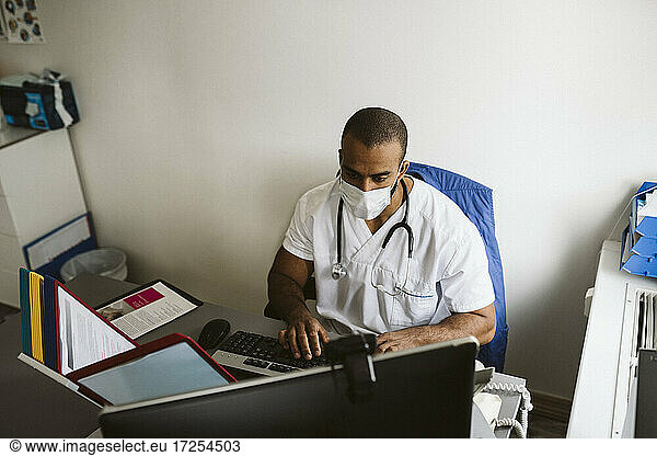 Male healthcare worker wearing face mask using computer in medical clinic