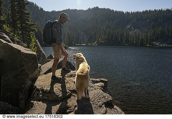Male hiker and dog standing on a cliff next to an alpine lake