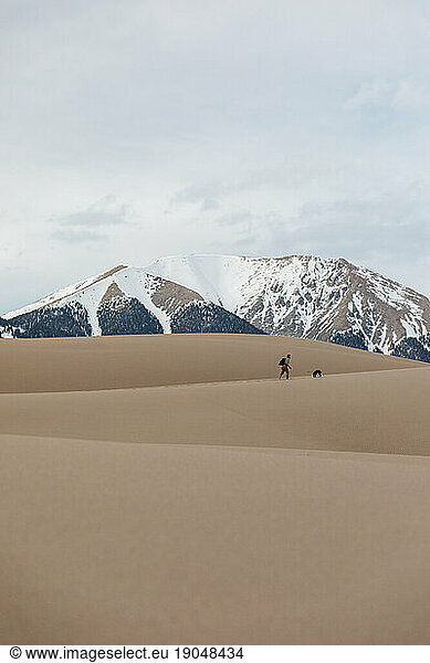 male hiker with puppy hike the layered sand dunes under snowy mountain