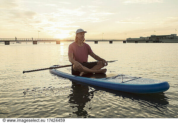 Male instructor meditating while sitting on floating paddleboard in sea