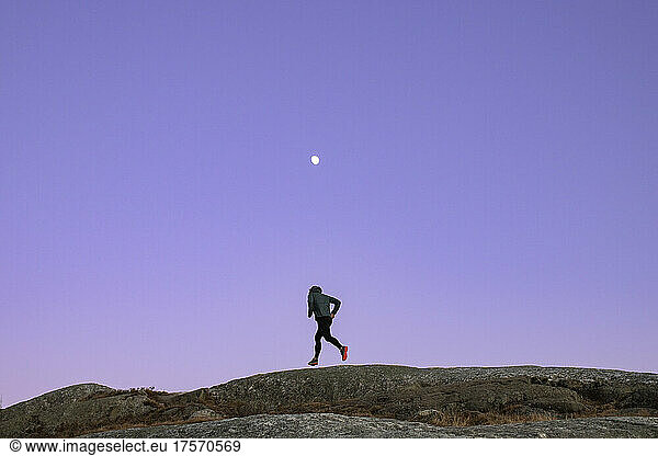 Male jogger runs along rocky summit at twilight with moon