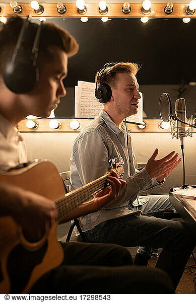 Male musicians recording vocal and guitar parts of song in professional studio