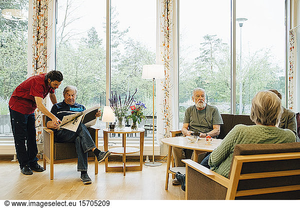 Male nurse standing by senior man reading newspaper while retired friends talking at nursing home