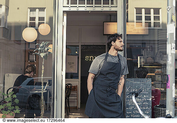Male owner looking away while standing with hands in pockets at cafe doorway