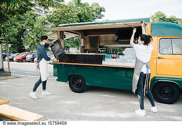 Male owner positioning board on concession stand while female coworker opening shade of food truck