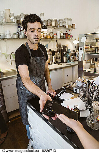 Male owner taking wireless payment from customer at checkout counter in cafe