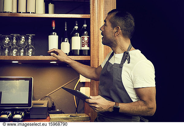Male owner with digital tablet looking at wine bottles while standing in cafe