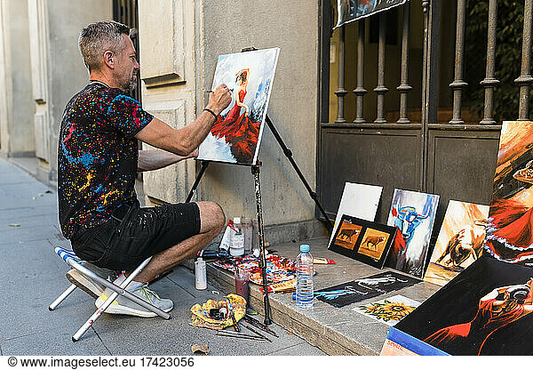 Male painter painting on canvas while sitting on street
