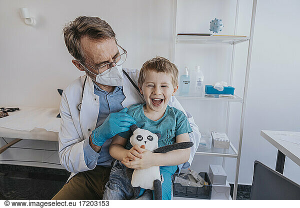 Male pediatrician looking at cheerful boy with toy while sitting at medical examination room