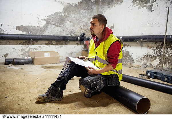 Male plumber reading document while sitting on floor by pipes at basement