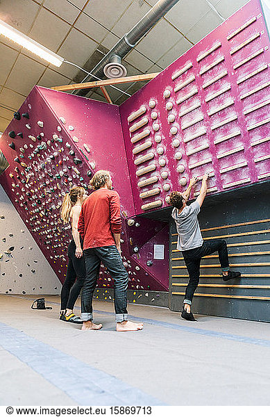 Male practicing wall climbing while mature coach and female student standing in gym