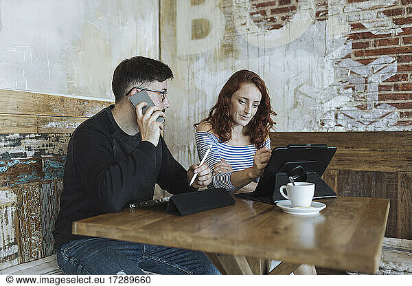 Male professional talking on smart phone while sitting with female colleague working at cafe
