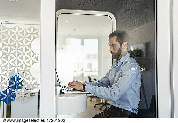 Male professional working on laptop while sitting in telephone booth at office