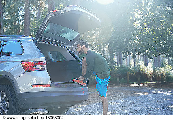 Male runner tying laces on car boot in sunlit forest