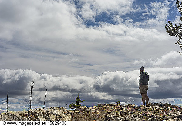 Male Standing On Mountain Top Looking At Cell Phone With Moody Clouds