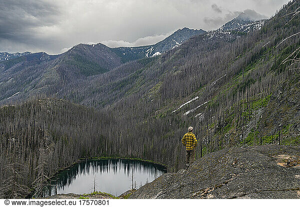 Male stands above alpine lake ravaged by wildfire