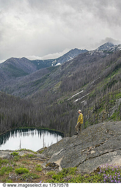 Male stands on a ledge above an alpine lake surrounded by burned trees