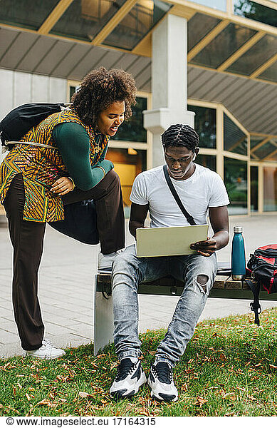 Male student e-learning on laptop while female friend standing by in campus