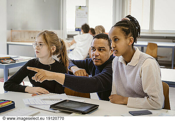 Male teacher pointing by female students sitting at desk in classroom