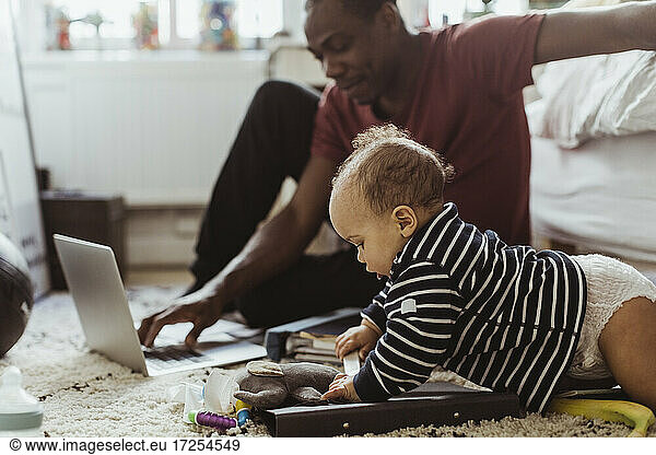 Male toddler playing with toys by father using laptop in bedroom