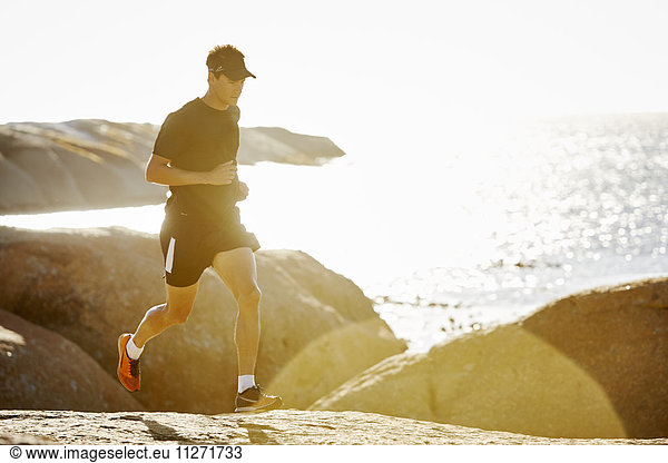 Male triathlete running on rocky trail along sunny ocean