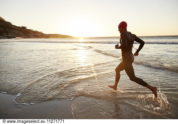 Male triathlete swimmer in wet suit running into ocean surf at sunrise