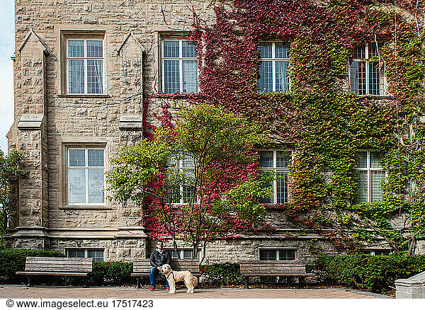 Man and dog sitting in front of red ivy covered building in fall.
