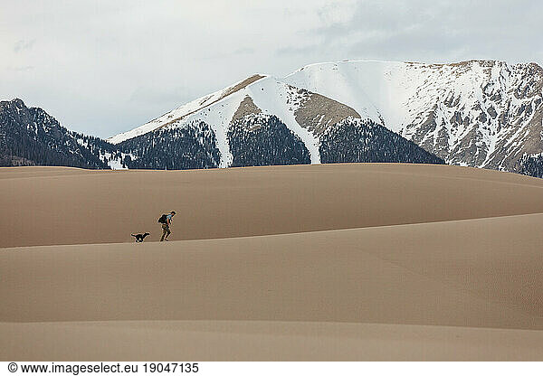 man and dog walk over sand dunes with snowy mountains in background