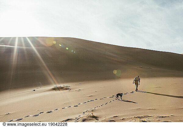 man and his dog walk up a giant sand dune in near sunset in colorado