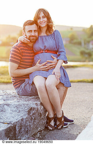 man and pregnant woman in dress are hugging in summer at sunset