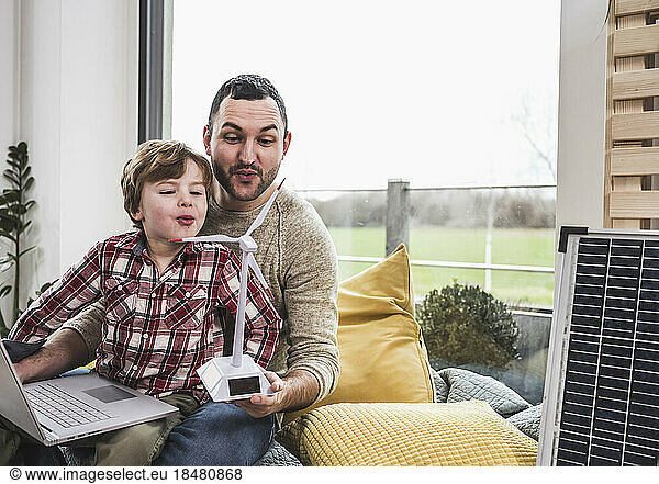 Man and son blowing on wind turbine model by cushions at home