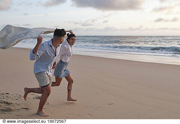 Man and woman running together holding blanket at beach